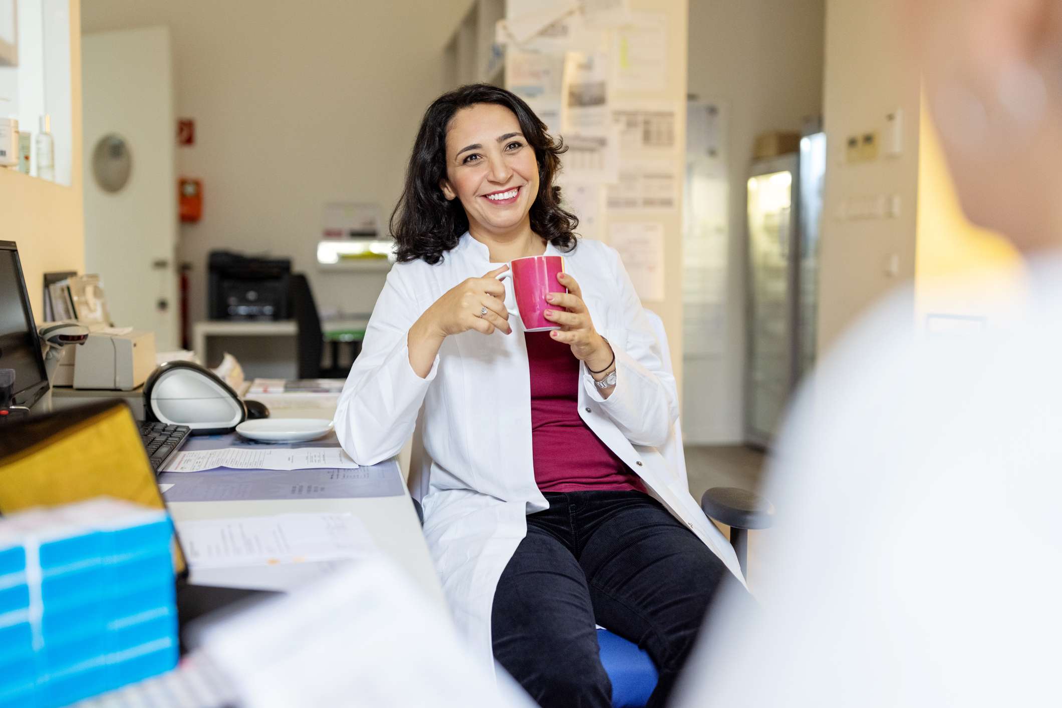 Female pharmacist sitting on chair and holding coffee, talking with female colleague during break