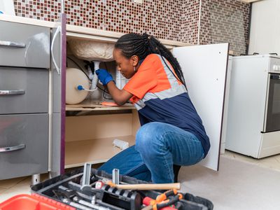 Black plumber in uniform sitting on the floor in kitchen and fixing pipe