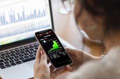 Person looking at a smartphone with a stock market index while sitting at a laptop showing other market data