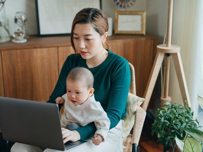 Woman holding a baby on her laptop while she works on a laptop at home. 