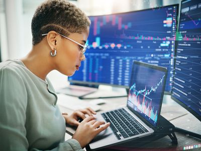 Female traders sitting at desk working on a laptop. Several monitors showing financial data are behind the laptop.