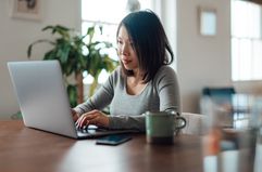 A woman sits at a table with a laptop and a mug