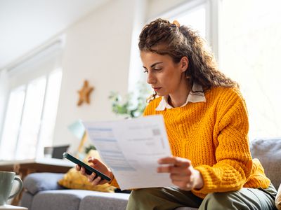 A woman with brown hair and a yellow sweater sits on a couch looking at a document and typing on her cell phone