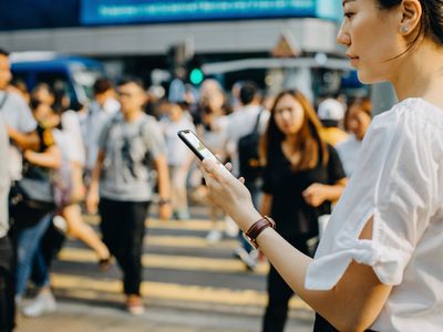 Young Asian woman checking financial trading data on smartphone in downtown city street, against busy pedestrian walkway and city traffic