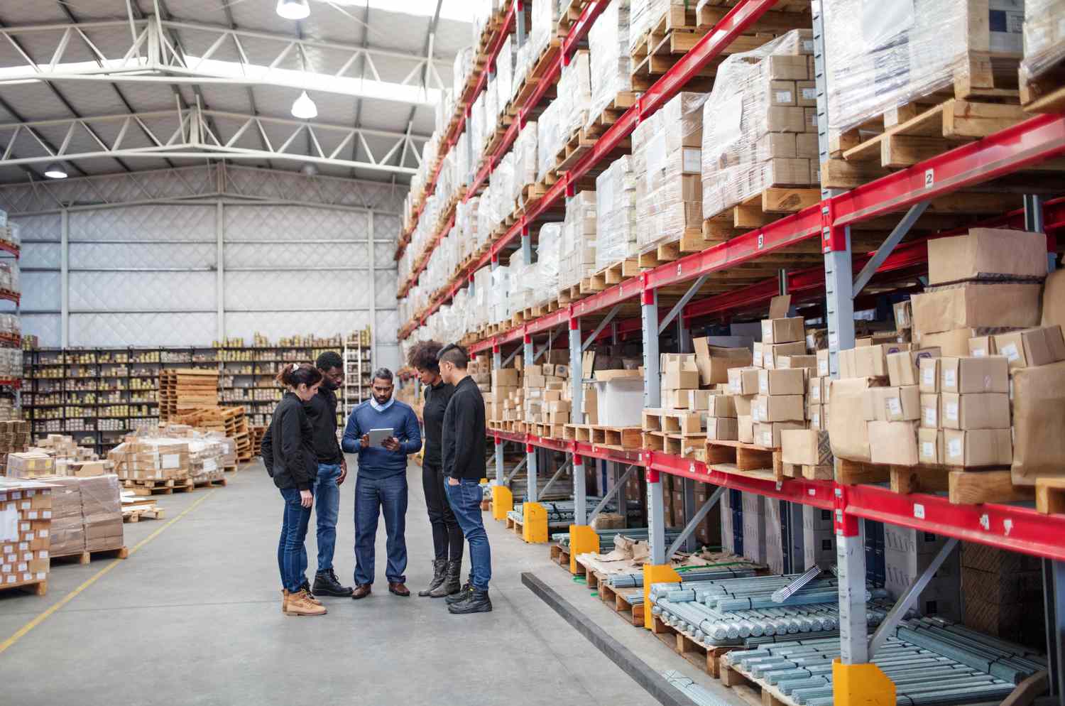 Group of people in a warehouse surrounding by boxes all looking at a tablet held by one person