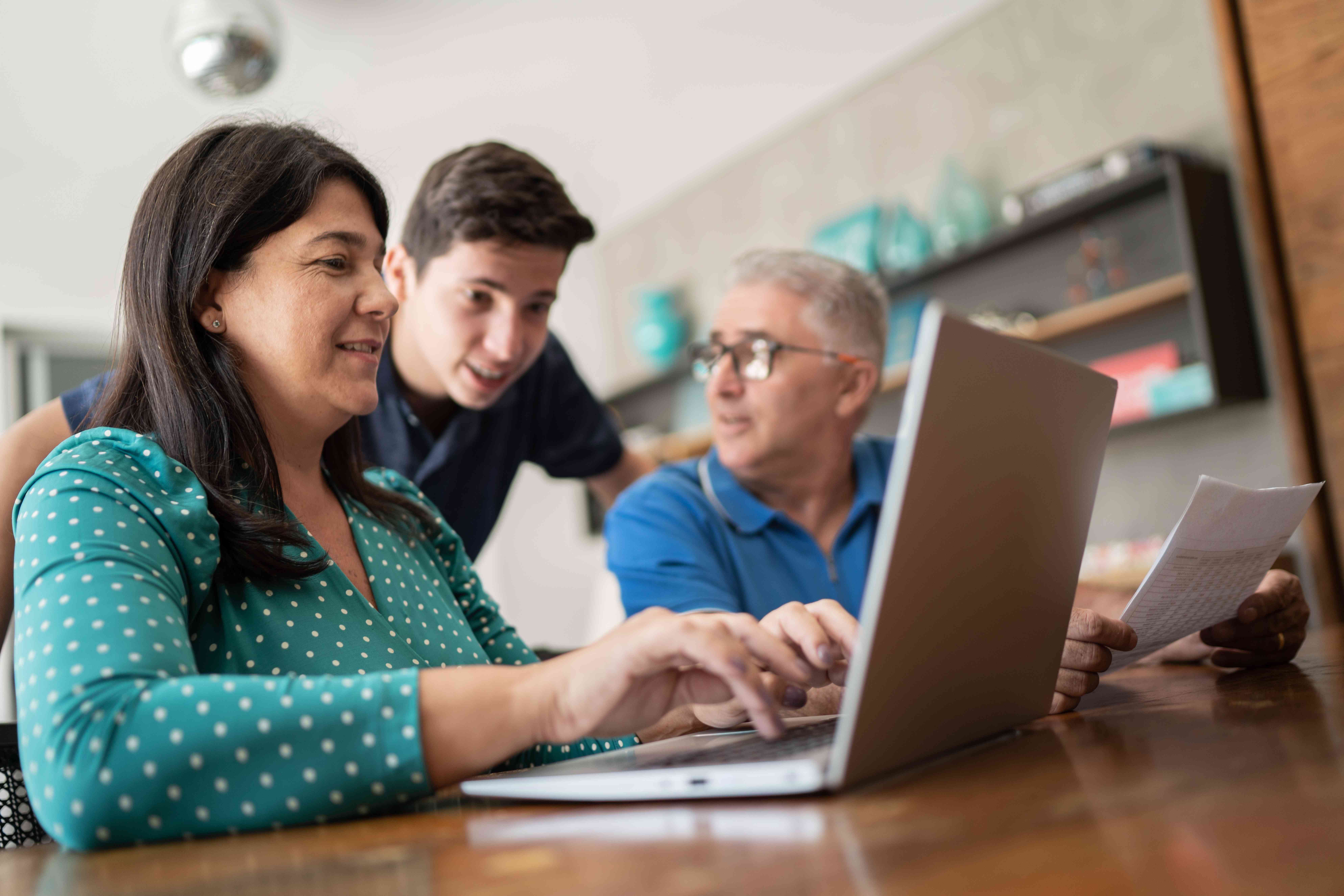 Parents sit at a table with a laptop while a teenaged child leans forward behind them as they explain the family investment account.