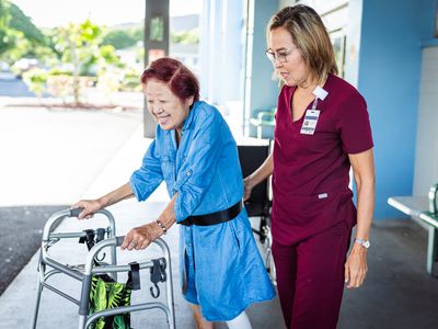 A caregiver helps her elderly patient walk with the help of a walker on a sidewalk