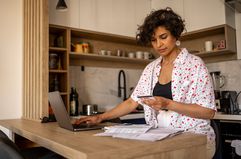 A woman stands in a kitchen at an island with a laptop and a credit card with documents sitting on the counter