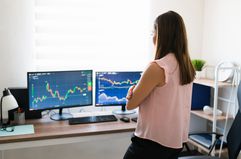 Standing woman looking at charts on two computer monitors