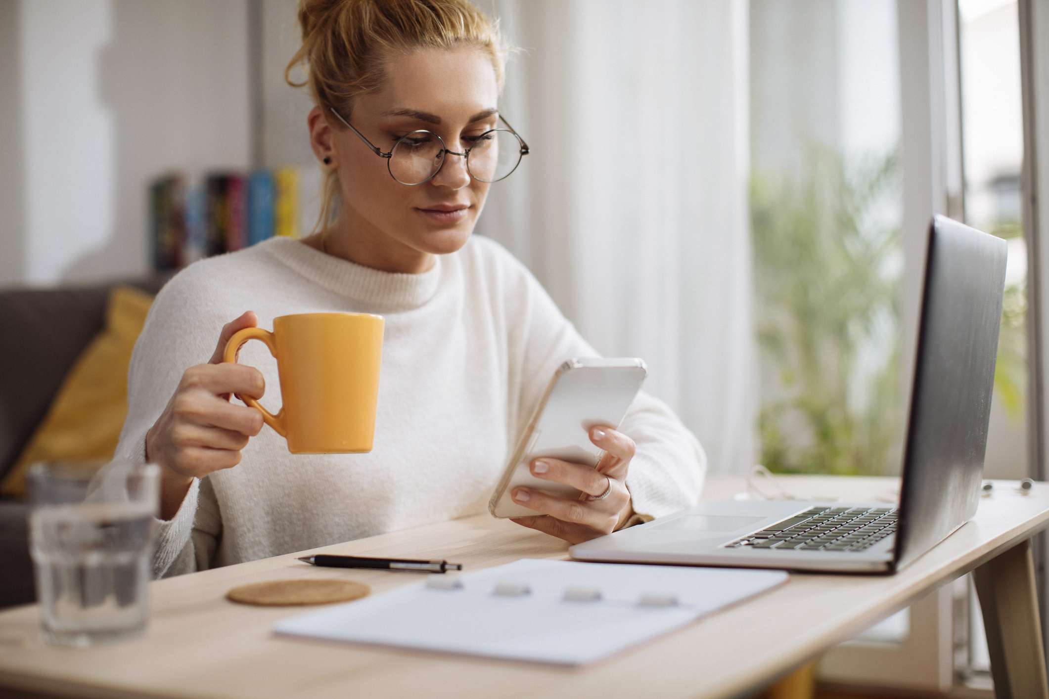 Young woman reading her phone while holding a cup of coffee. She sits in a home office in front of a laptop with a notepad and pen. 