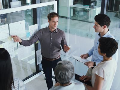 A group of office workers standing in a semi circle as one man points at a roller white board with papers taped to it, shot from above. 