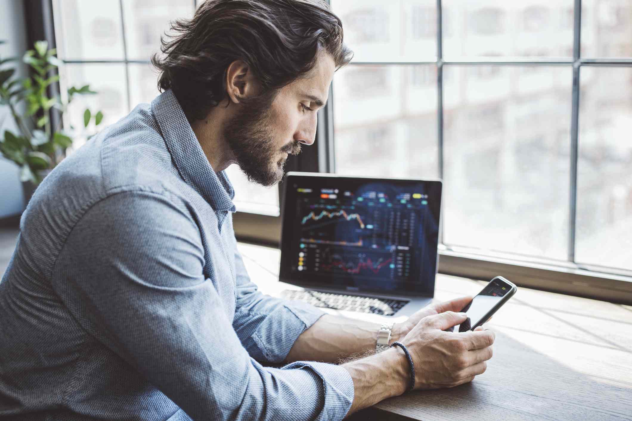 Young man sitting at a counter with a laptop showing market data and holding a smartphone