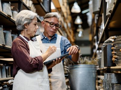 Two older adults with a table and stylus standing in an aisle with equipment and products all around them. 