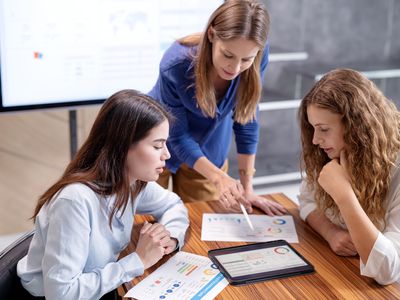 Three women in a conference room. One standing over them discussing a document.