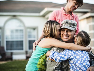 Mother watching over two daughters hugging their dad in military fatigues uniform in the front yard of their house.