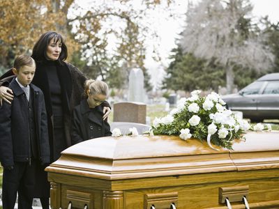 A widow stands with their arms around two young children next to a casket at a cemetery.