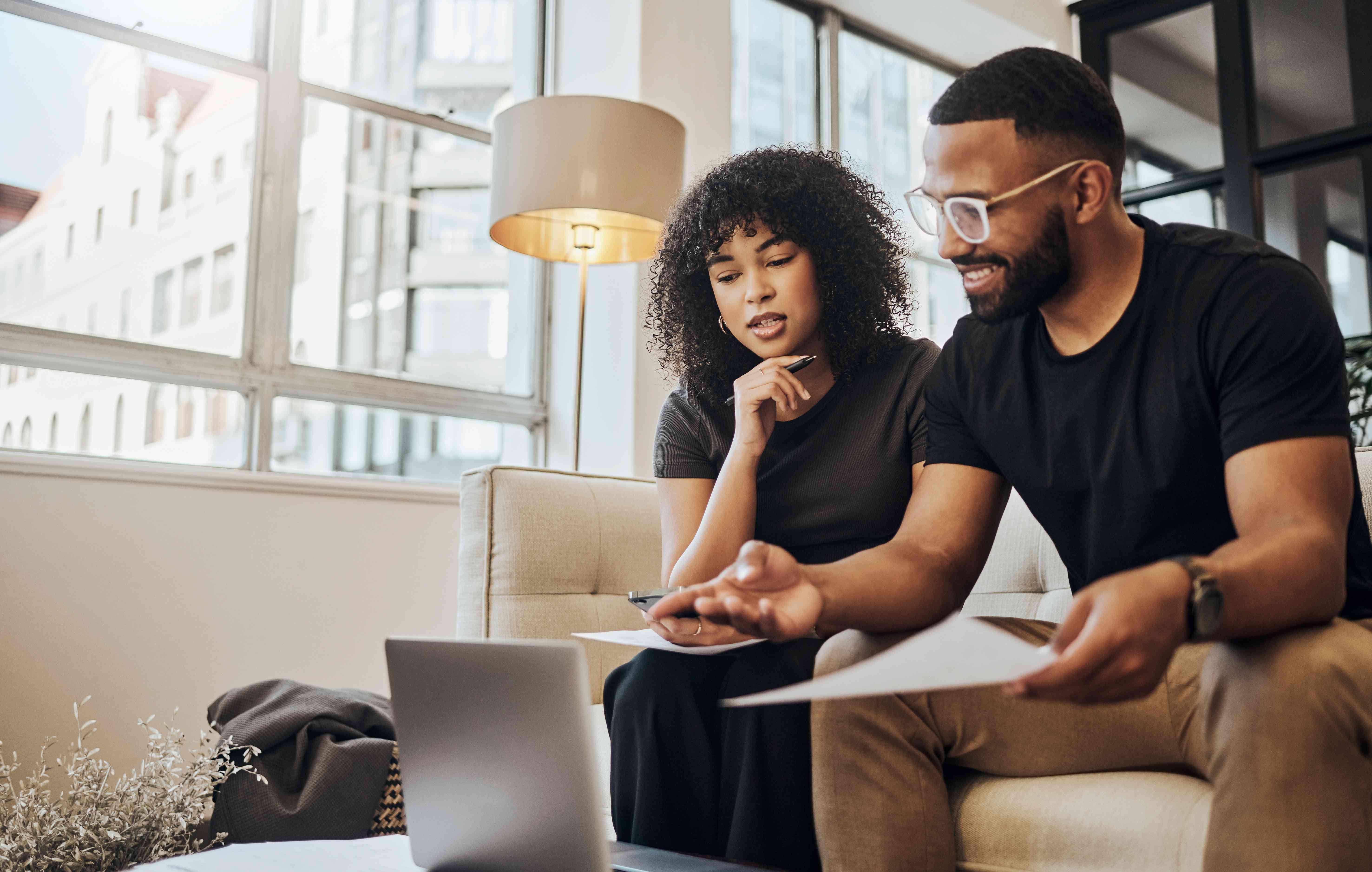 A couple sits on a couch holding documents and looking at a laptop; they're opening a business bank account.