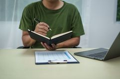 A man writing in an open notebook with an open laptop on the table