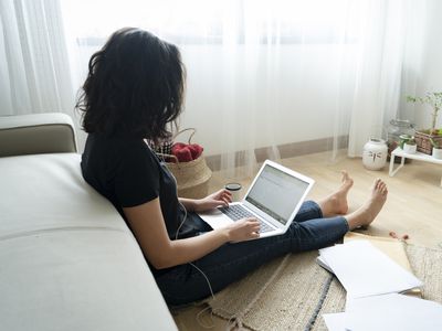 A woman in a black shirt and jeans sits barefoot on the floor with a laptop and paperwork