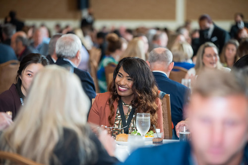 A woman with long, wavy hair and a bright smile sits at a round table during a formal event, surrounded by other attendees. She is wearing a brown cardigan and a patterned blouse.