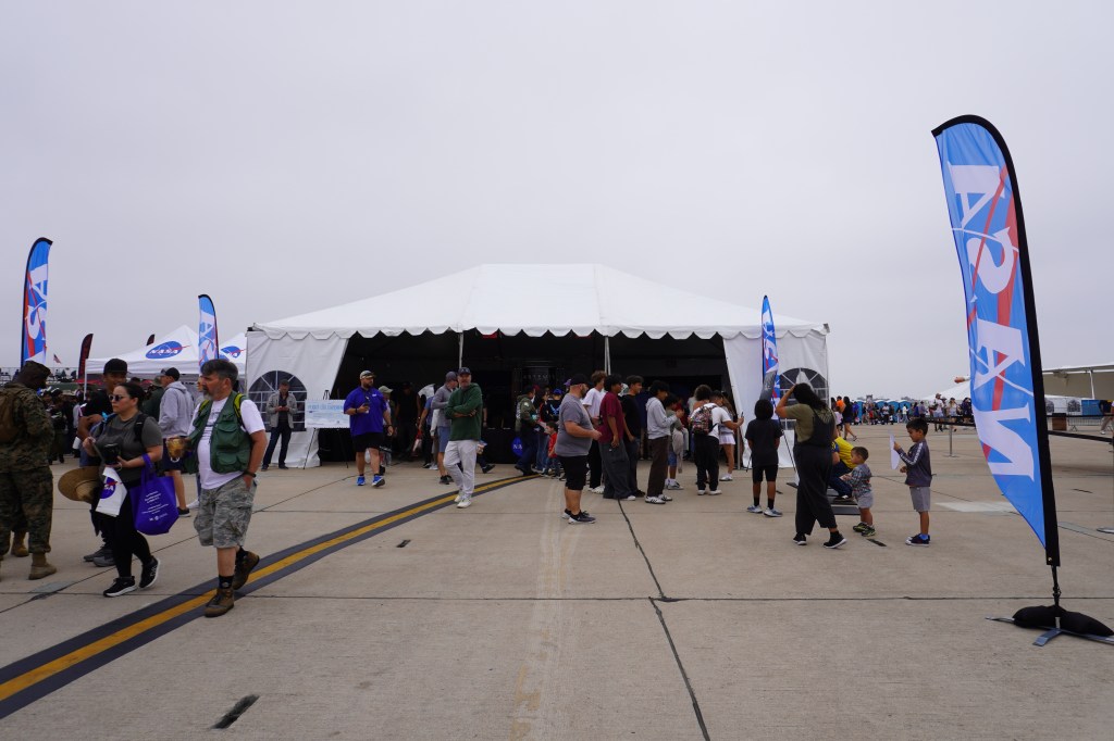 A view of the front of a bustling event tent outfitted with NASA flags and insignia. The tent is centered in the photo and sits atop a concrete airfield, where many people are walking in and out of the tent. A grey-white overcast sky is behind and above the tent.