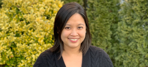 Image: A woman with dark hair and dark eyes in a cable-knit v-neck sweater smiles into the camera. She is pictured against a background of yellow and green trees.