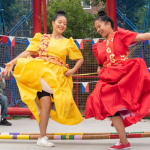 Two dancers in center stage, dressed vibrantly, with musicians in the background and additional performers on either side