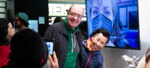 Two individuals smile, posing for a photographer in a gallery setting