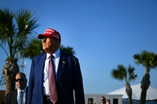 President-elect Donald Trump, wearing a suit and a Make America Great Again ball cap in an outdoor setting with palm trees