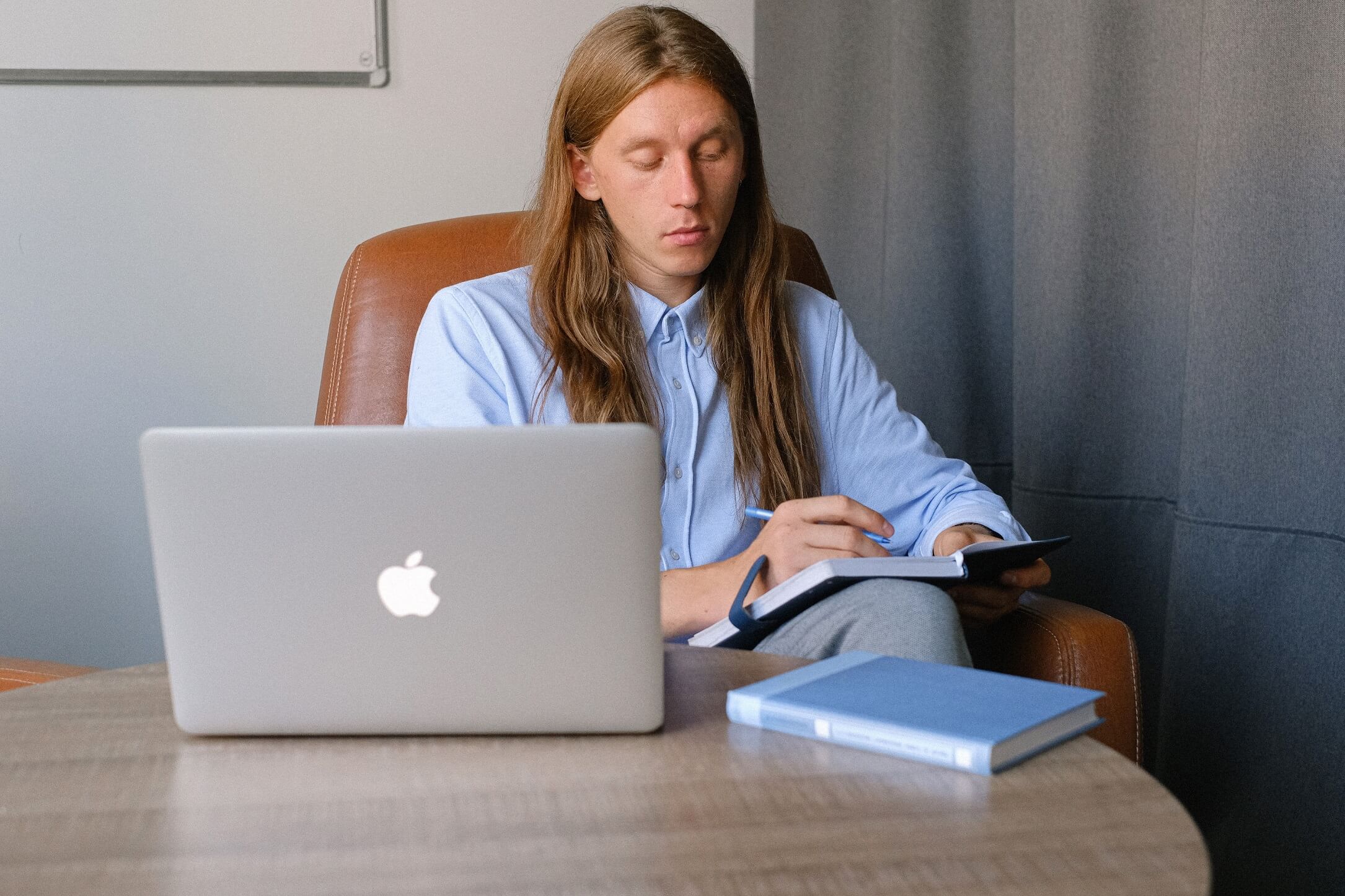 A man a sitting on a chair and wiriting in a notebook. There is a laptop on the table in front of him.