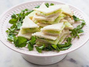 Triangle cut cucumber tea sandwiches are stacked on a cake stand and encircled with fresh herbs. Sliced cucucumbers are visible inside each one.