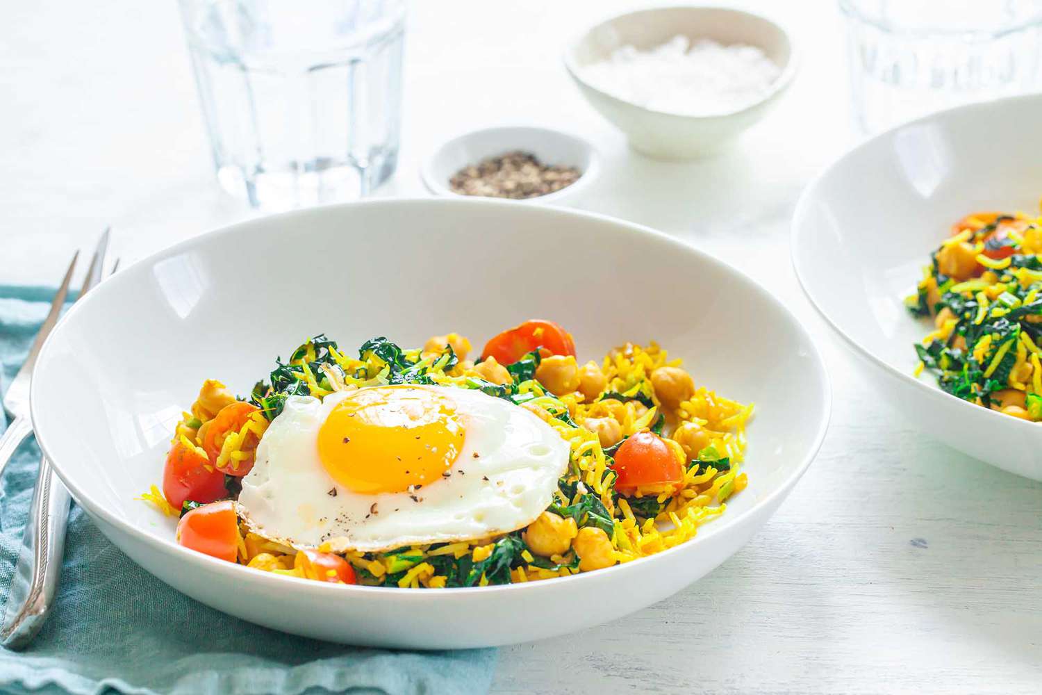 Side view of a bowl of turmeric fried rice with greens topped with a fried egg. Chopped kale, tomatores and chickpeas are mixed in with the rice. Silverware and a linen are to the left. A second bowl of turmeric fried rice with greens is in partial view to the right.