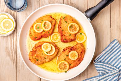 Italian butter chicken (petti di pollo al burro) in a skillet at a table with a plate of lemon slices, a glass of water, and a table napkin