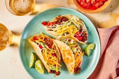 Crispy ground beef tacos on a plate at a table setting with glasses of beer, bowl of diced tomatoes, and a table napkin