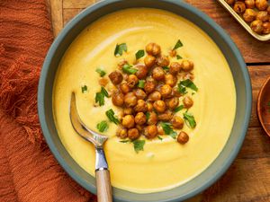 Bowls of hummus next to a small bowl of roasted chickpeas, another small bowl with chopped parsley, and a burnt orange kitchen towel, all on a wooden table