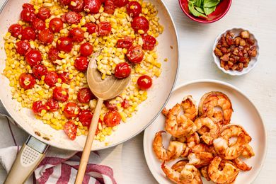 Skillet with roasted tomatoes and corn in a cream sauce next to a plate of stir-fried shrimp, a small bowl of pancetta, and a small bowl of basil leaves, all sitting on the counter