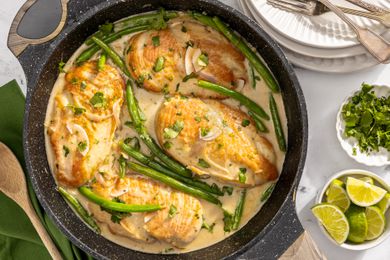 creamy coconut lime chicken in a pan at a table setting with a stack of plates, a bowl of minced cilantro, and a bowl of lime wedges