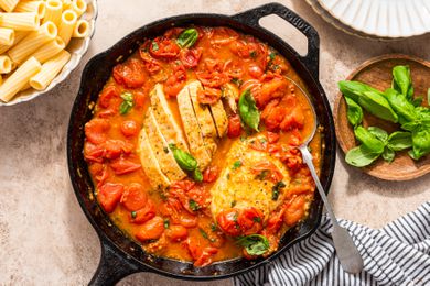 chicken pomodoro in a cast iron skillet at a table setting with a bowl of rigatoni, bowl of basil, stack of plates, and a table napkin