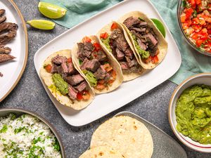 Platter of Arrachera (Mexican Skirt Steak) Tacos With Lime Wedges, and in the Surroundings, a Platter of Arrachera (Mexican Skirt Steak), a Bowl of Cilantro Rice, a Bowl of Salsa, a Bowl of Guacamole, and a Tray of Tortillas, All on a Light Teal Kitchen Towel