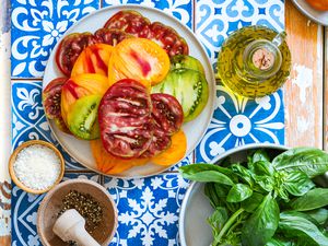 sliced heirloom tomatoes on a plate next to a bowl of coarse salt, crushed pepper in a mortar and pestle, carafe with olive oil, and a bowl of fresh basil, all on a spanish-tile covered tray