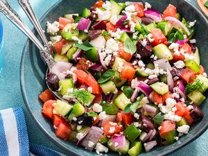 Bowl of Easy Greek Salad With Utensils, and Next to It, a Glass of Water, a Small Bowl of Herbs, and a Kitchen Towel