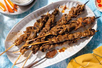 Sach Ko Jakak (Cambodian Grilled Lemongrass Beef Skewers) in a Platter Surrounded by a Plate With Sliced Carrot and Daikon Radish Pickle and a Bowl of Rice and a Yellow Kitchen Towel Next to It