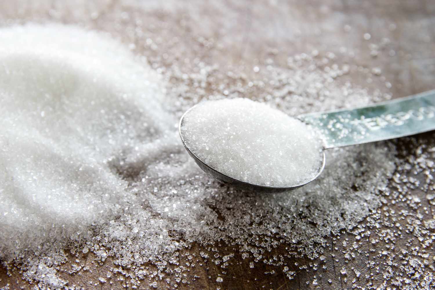 A teaspoon full of sugar with a pile of sugar next to it on a wood surface