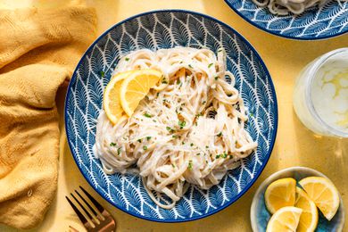 creamy lemon riccotta pasta in a bowl surrounded by a table napkin, a glass of water, and a bowl of sliced lemons