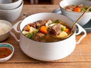 Ladle raising some nilagang baka from a Dutch oven with more, and in the surroundings, a stack of bowls, a serving of Filipino beef soup in a bowl, a bowl of rice, and a small bowl of fish sauce