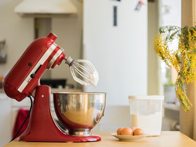 Side view of a red stand mixer and metal bowl with the top section tilted back showing the whisk attachment. The mixer is on a kitchen counter next to a plate of eggs and container of flour