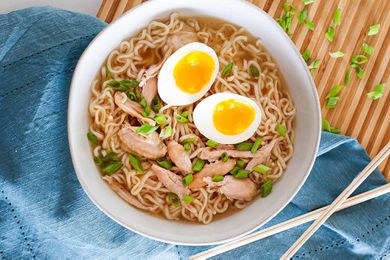 A white bowl, sitting next to a blue napkin on a cutting board. The bowl is filled with the best chicken wing ramen. It has noodles, chicken, scallions, and a soft boiled egg -- all made in the pressure cooker.