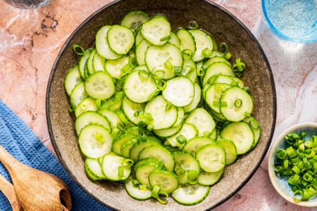 cucumber salad in a bowl at a table setting with a bowl of slcied scallions, serving utensils on a table napkin, and a glass of water 