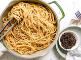 Pot of Vegan Cacio e Pepe with Some Noodles Wrapped around a Pair of Tongs Next to a Bowl of Black Pepper on a Small Plate and Two Forks on the Counter