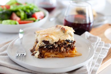 A piece of eggplant moussaka on a plate with a glass of wine and a salad behind it.